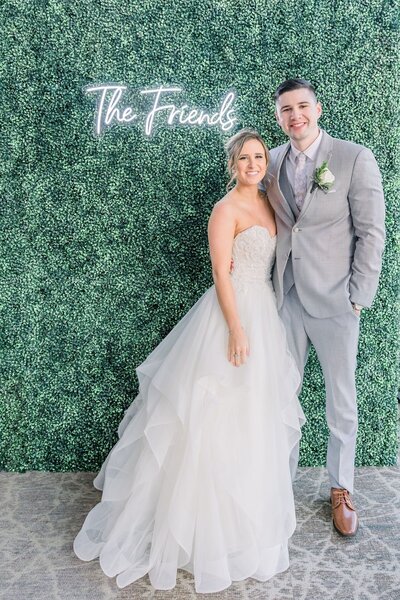 Bride with a fluffy gown standing next to groom in a tall suite in front of a leafy wall with a neon sign that reads "The Friends"