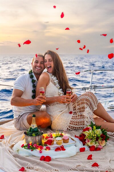 Engaged couple celebrating on a sailboat in Hawaii as rose petals fall around them, holding drinks and smiling during their post-proposal moment.
