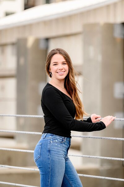 A senior grad in a black long-sleeve shirt and blue jeans leans against a railing outdoors, smiling at the camera.