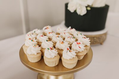 Photo of a gold cake stand with vanilla mini cupcakes, topped with white buttercream and rainbow sprinkles.