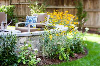 A stunning backyard transformation featuring a curved stone seating area surrounded by lush greenery, vibrant yellow flowers, and cozy outdoor cushions.