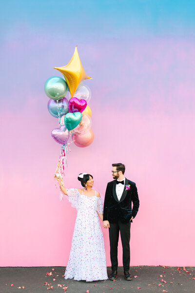 wedding couple holding balloons standing against pastel wall