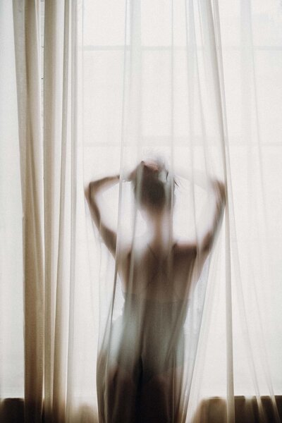 Silhouette de femme en body de dos, derrière un rideau léger