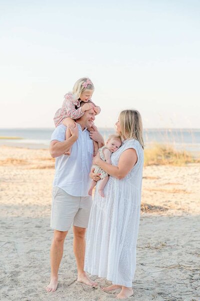 Toddler girl on dad's shoulders while dad lovingly gazes at mom and baby at Fish Haul Beach Park in Hilton Head, SC with Hilton Head Photographer, Lamp and Light Photography.