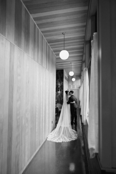 Groom kissing the bride on the cheek while standing in a long corridor.