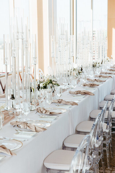 This image features a sophisticated arrangement of three white long tables set in Venue TwentyTwo, complete with acrylic chairs and elegant glass candelabras. Each table is adorned with beautiful floral centerpieces, charger plates, and taupe knotted napkins, creating a chic and modern atmosphere. Ideal for couples looking for unique wedding decor ideas, this setup showcases how to elevate any reception with stylish details in Ottawa.