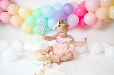 Little girl is sitting on a white backdrop with a pastel rainbow balloon garland in the background. She is smiling excitedly down at her smash cake while extending her arms out and lifting one foot. She wears a pink dress that has "one" spelled out in cursive.