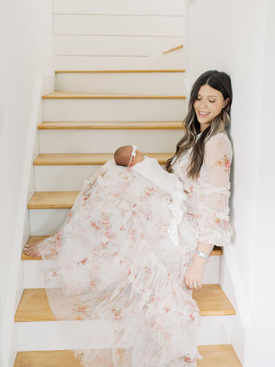 a mother sits on the stairs in her home holding her newborn baby in her lap, she is wearing a breautiful spring flowing dress