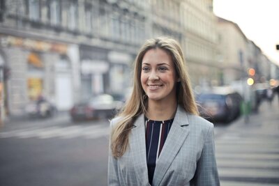 A smiling woman with long blonde hair wearing a plaid blazer and striped shirt, walking down a city street with blurred buildings and cars in the background.