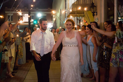 A bride and groom walk hand in hand outside at night, smiling as guests on both sides hold sparklers and cheer, creating a festive tunnel for their wedding celebration.