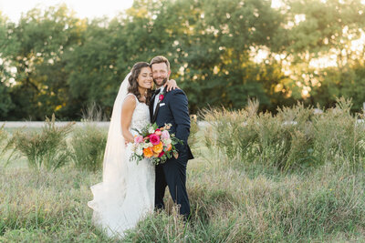 wedding couple hugging and looking at camera standing in a field with sunset behind them
