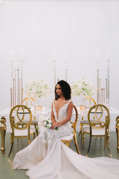 Bride holds wedding bouquet and sits at wedding reception table in gold chair