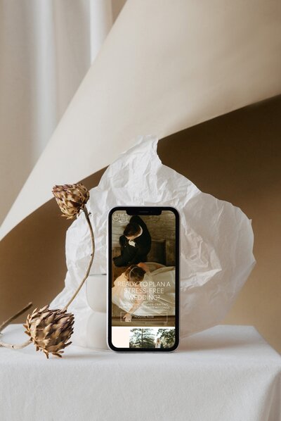 A smartphone displaying a wedding planning website, meticulously crafted through custom Showit website design, rests against a white cloth backdrop, alongside two dried flowers on a pristine white surface.