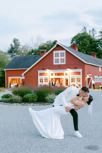 groom in white dinner jacket dips bride in front of red barn wedding venue