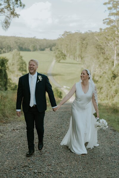 A joyful and heartfelt wedding photo of a bride and groom in Bonville walking up a scenic hill holding hands and laughing