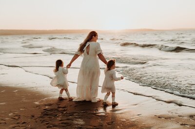 A mother in a long white dress holds the hands of her 2 young duaghters, also in white, as they walk along the shore