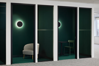 A small phone room featuring a comfortable, modern beige chair, soft lighting from a wall sconce, and forest green walls and carpet