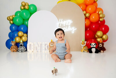 Boy in a blue romper sitting near beige and white balloon arches, surrounded by stuffed jungle animals.