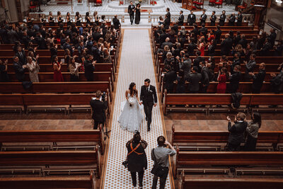 Kristen stands in a large wedding chapel taking a picture of the bride and groom exciting their ceremony