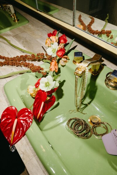 A green bathroom sink with a modern red and orange wedding bouquet and gold jewelry strewn around.
