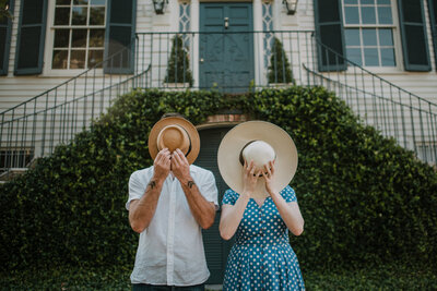 two people holding hats in front of their faces with house in the background