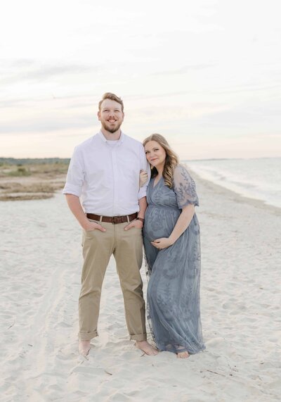 Bluffton Maternity Photographer, Lamp and Light Photography captures a pregnant woman and her husband standing on the beach and smiling lovingly at sunset.