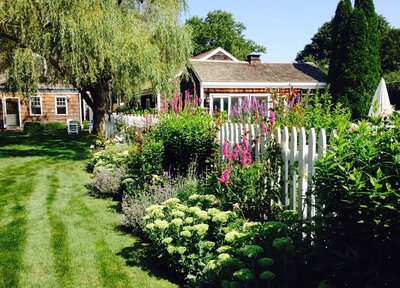 Vibrant garden with lush flowering plants and greenery growing along a white picket fence, leading to a charming cottage-style home in the background.