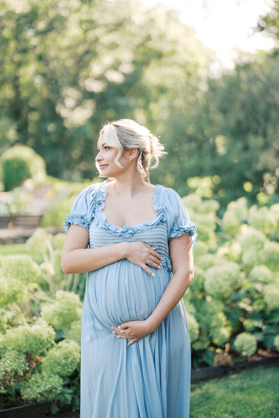 Woman with brown hair wearing a pink maternity gown stands on beach looking away holding her pregnant belly.