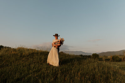 A woman in a white skirt and dark top with her fiancé wearing a cowboy hat on dirt path in a field, entangled in one another's love. The sun is low, casting golden light. The field surround the path if green, with blue sky above. The setting is a serene outdoor setting—perfect for capturing moments by a New England Portrait Photographer.
