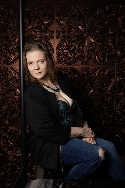 Woman in jeans and a black jacket and teal shirt and matching jewelry sitting on a stool in front of a decorative copper backdrop.