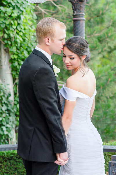 A groom in a black suit kisses his bride on the forehead as they stand on the grounds of Sleepy Ridge Weddings in the summer
