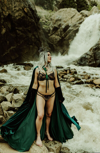 A woman in black lingerie with a black lace veil  smelling a pink peony in front of a roaring river for boudoir photos in Boulder Colorado by Kendra Colleen Photography