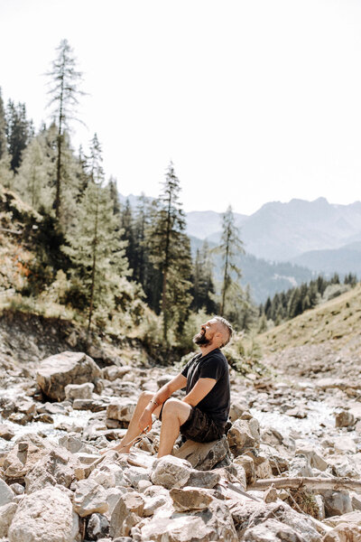 Alec smiling into the sky while sitting on a  large rock in a river bed, surrounded by mountains.