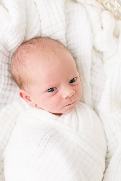 newborn baby wrapped in white awake and staring at the camera. Laying on a white blanket.