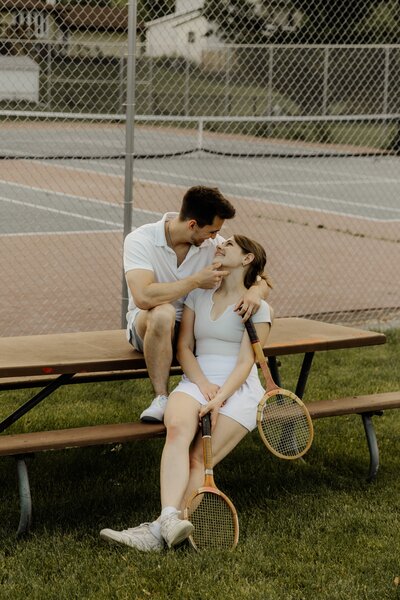 A couple sits on a picnic table with tennis rackets in hand, faces pulled in close.