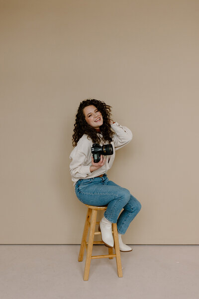 woman sits on stool holding a camera smiling