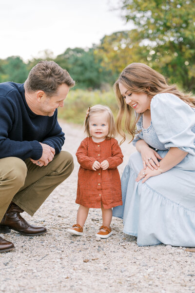 Mom in blue dress and dad in navy sweater squat next to toddler girl in rust dress during golden hour family photo session