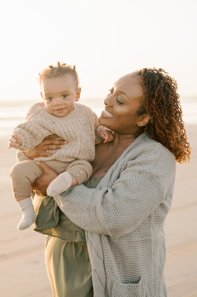 mother holding young baby boy on the beach in neutral colored knit outfit