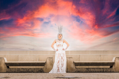 senior in a white snow queen avant garde outfit with crown on a rooftop in kansas city mo with pink clouds during sunset