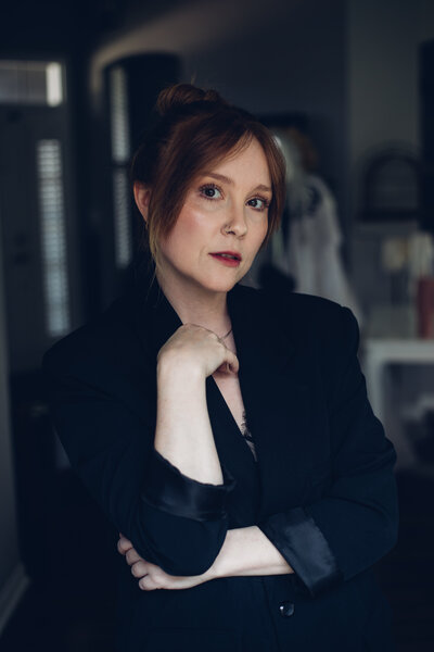 A woman with red hair in a bun, wearing a dark blazer, stands indoors with her hand resting near her face. She gazes directly at the camera as the owner of an agency providing branding and web design for female entrepreneurs.