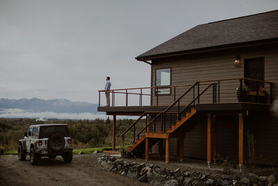 Groom stands on balcony overlooking the mountains in Alaska