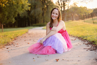HS Senior in a multi-colored pink, light pink, lavender and pink tiered dress sitting on a concrete path with autumn leaves scattered around in beautiful backlit sunset.