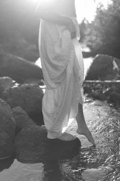 woman in a long white dress dips her toes in a river which glistens in the sun