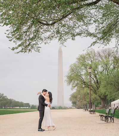 An Asian engaged couple sharing a kiss under a tree in front of the Washington Monument enjoying their Raleigh wedding photos by JoLynn Photography