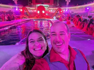 A couple in dressy attire poses with big smiles in front of a pool on a cruise ship deck. Behind the pool, a large inflatable red octopus is sprawled, its eyes lit from within and tentacles raised.