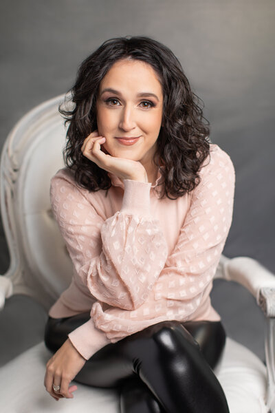 A woman with black curly hair wearing a pink top sits on a white chair in front of a gray backdrop
