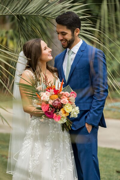 woman in white dress with bouquet smiling at man in blue suit in front of palm leaves 