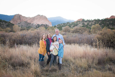 A happy family stands together smiling in tall golden grass with small white dog