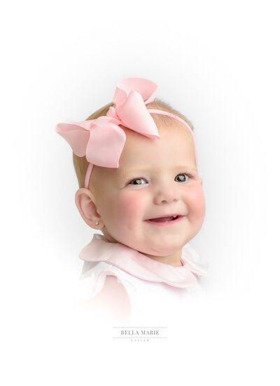 Baby girl with head turned to shoulder with a big smile, white shirt and pink bow.