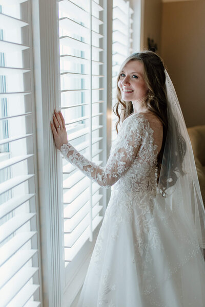 candid portrait of bride in white lace Stella York wedding dress and cathedral veil looking out window before Harelton, TX church wedding